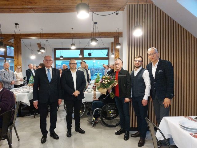 Kilka osób stoi, w tle sala, stoły. 